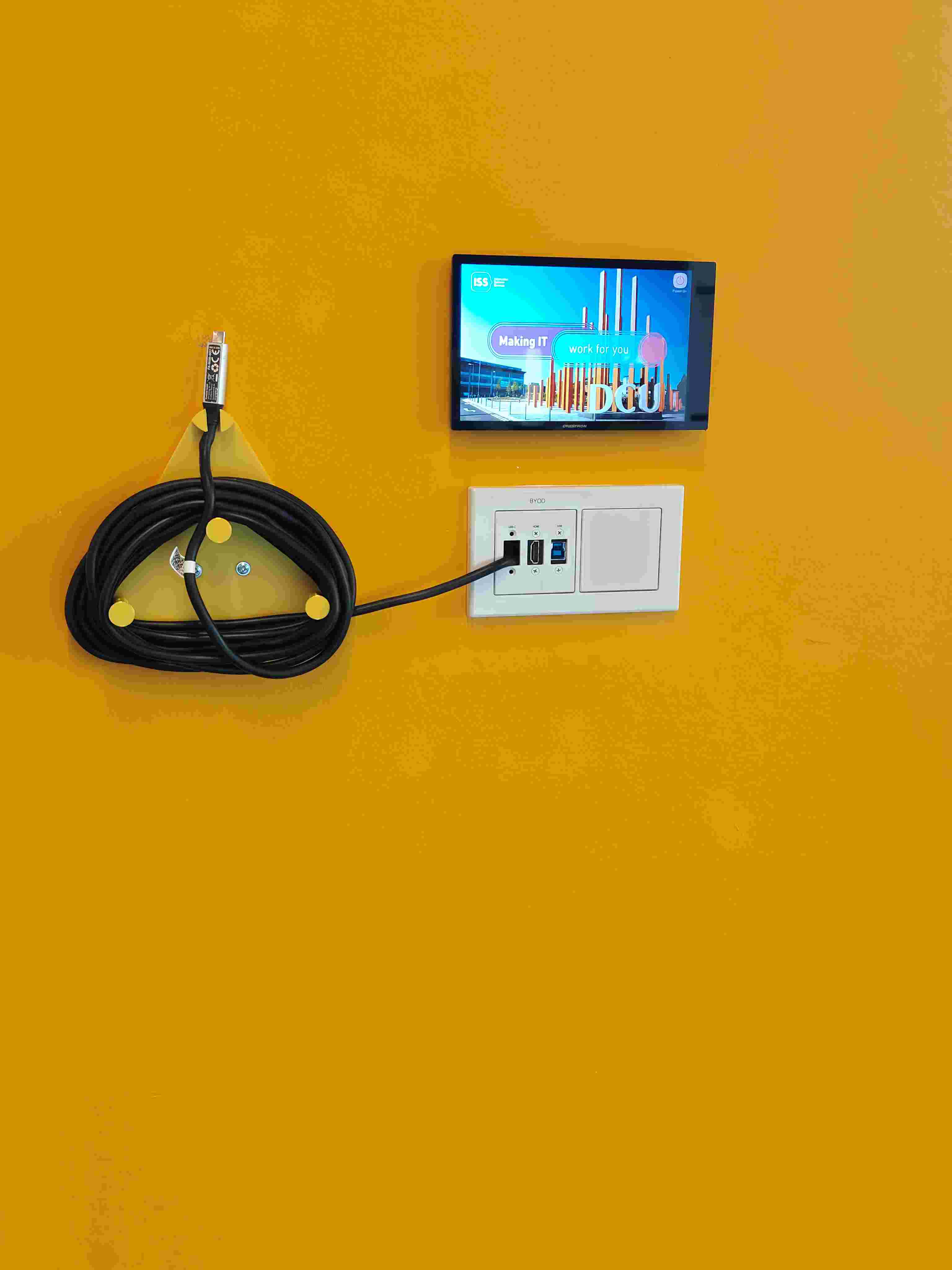 Yellow wall-mounted BYOD panel with a triangular cable wrap holder — the existing setup before I designed the dedicated projector cable hanger.