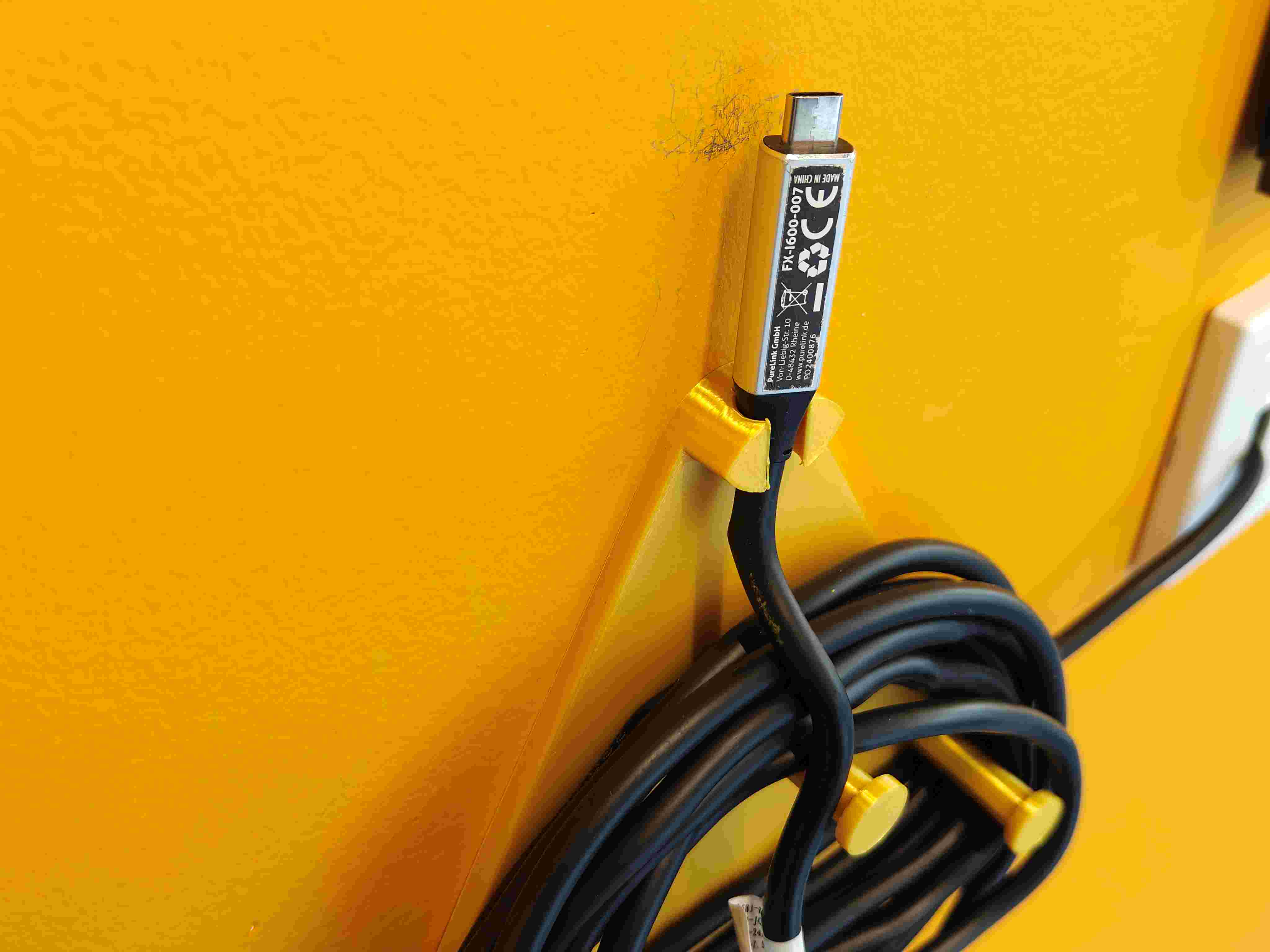 3D-printed cable hanger on a yellow wall, holding a PureLink display cable by the connector to prevent cable jacket wear and trip hazards.