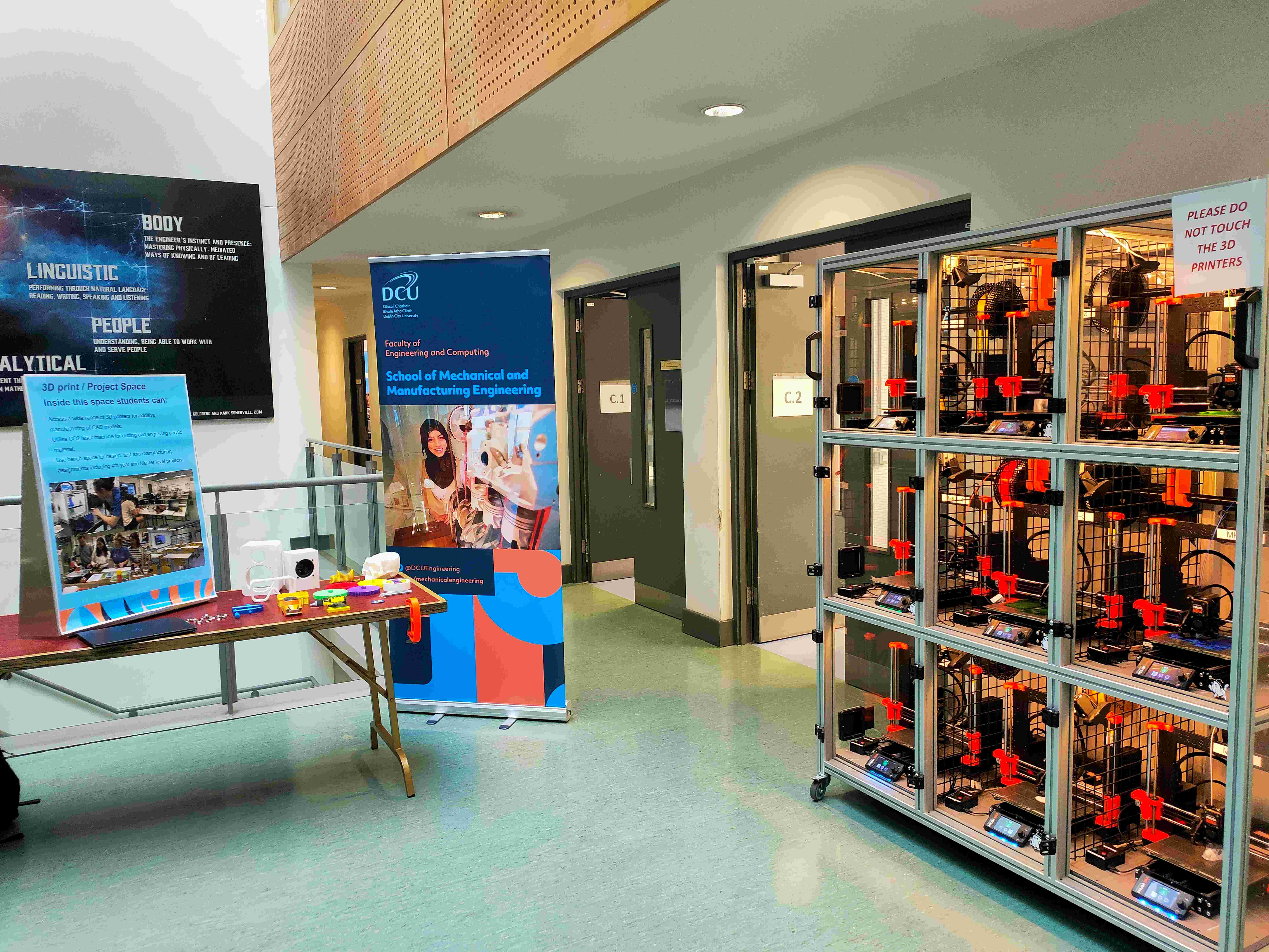 Open day exhibition booth — 3D print sample table with posters, DCU Engineering banner, and the MK4 print rack visible behind with a DO NOT TOUCH sign.