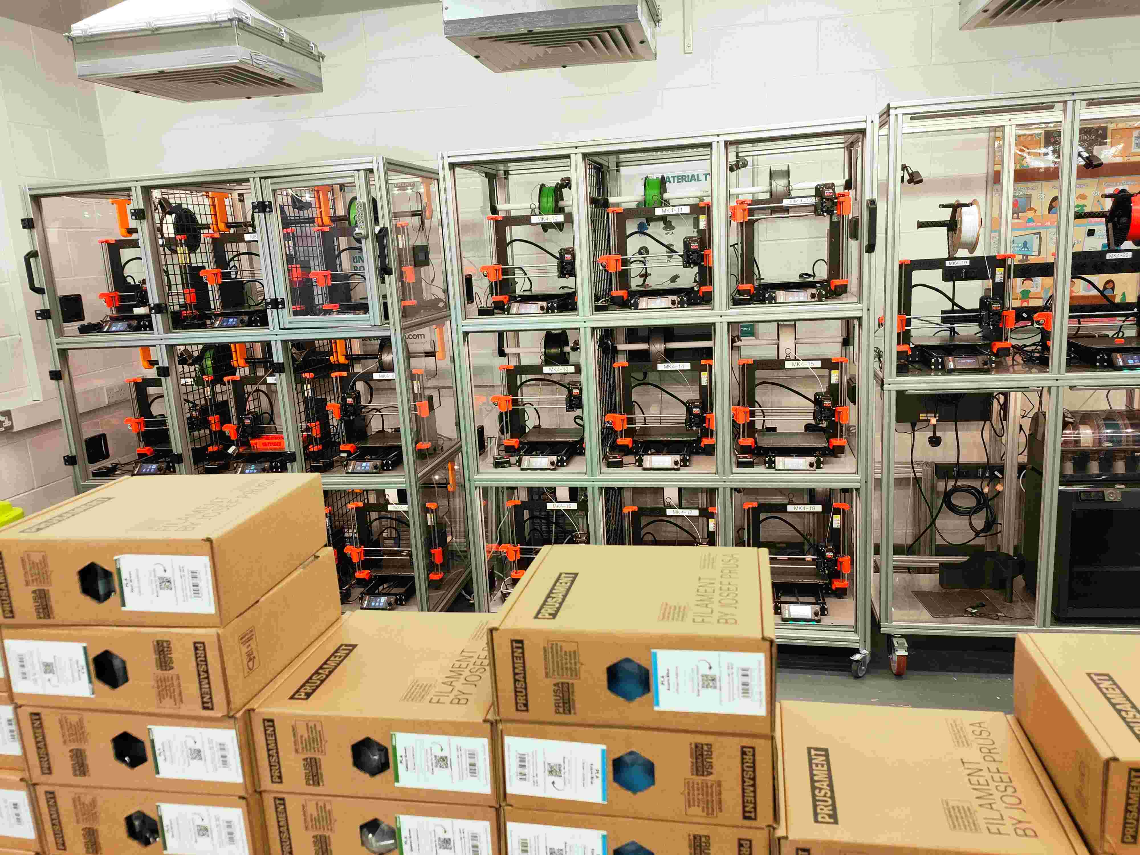 Prusa MK4 print farm with floor-to-ceiling Prusament filament stock, each printer bay showing machine ID tags MK4-10 through MK4-13.